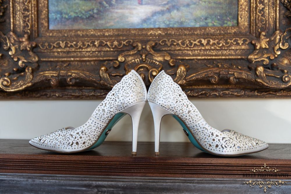 Elegant white crystal-studded wedding heels displayed in front of ornate gold frame.