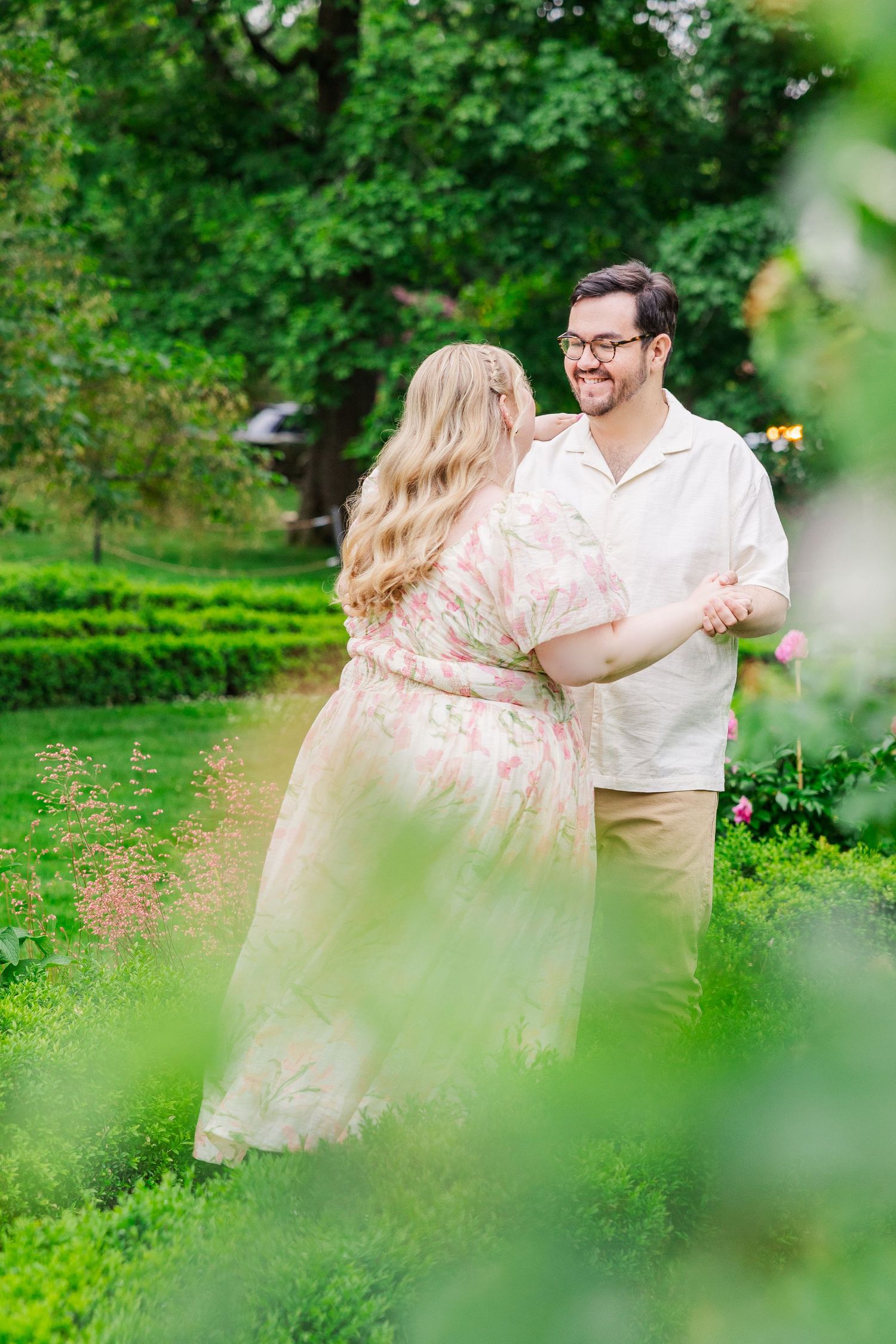 A couple embraces in a lush garden setting surrounded by green foliage on a sunny day.