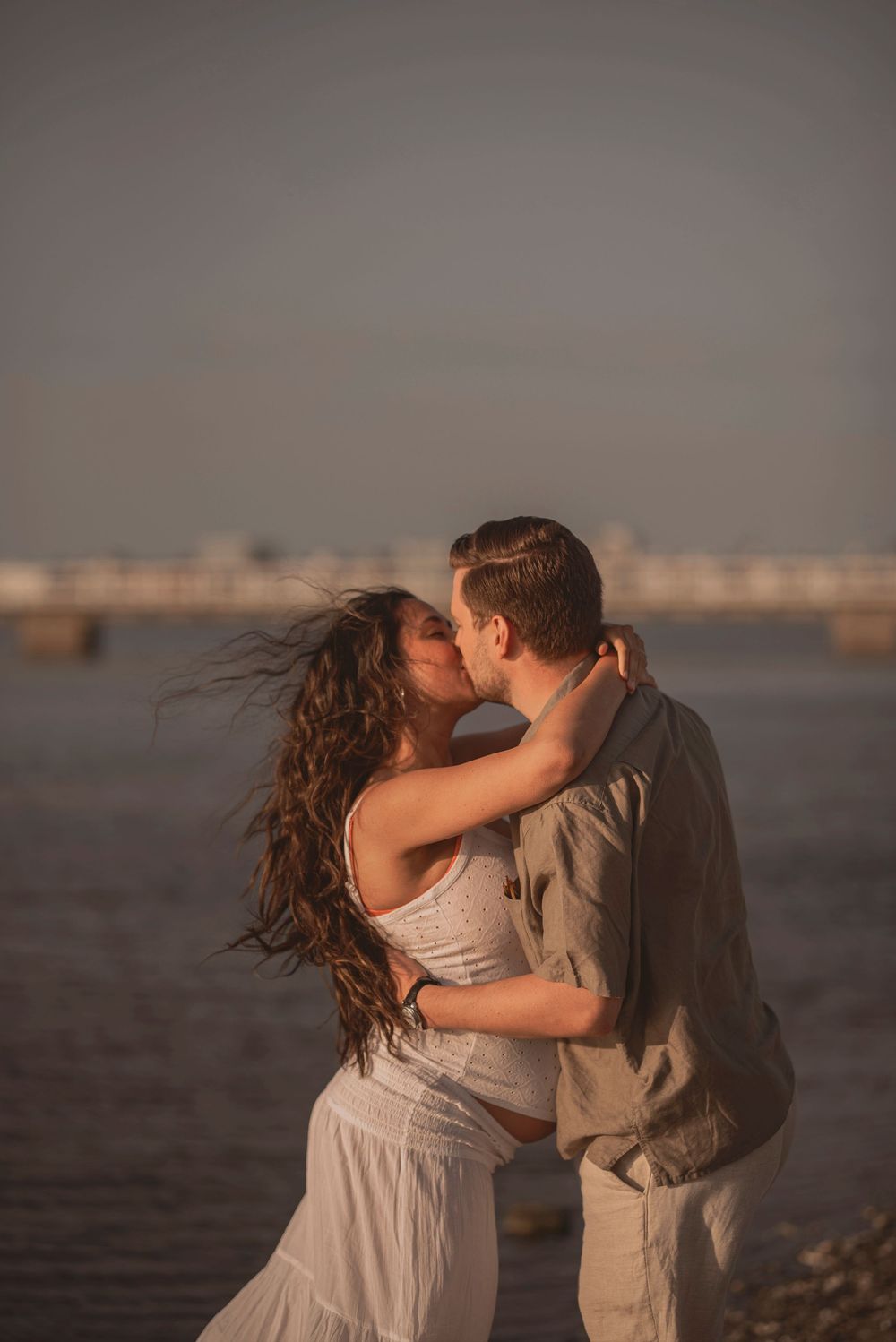 Silhouetted couple shares a romantic kiss at sunset with the bride's flowing dress and hair caught in the breeze.