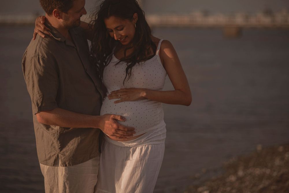 A couple stands together at sunset by the water's edge, lovingly cradling a pregnant belly.