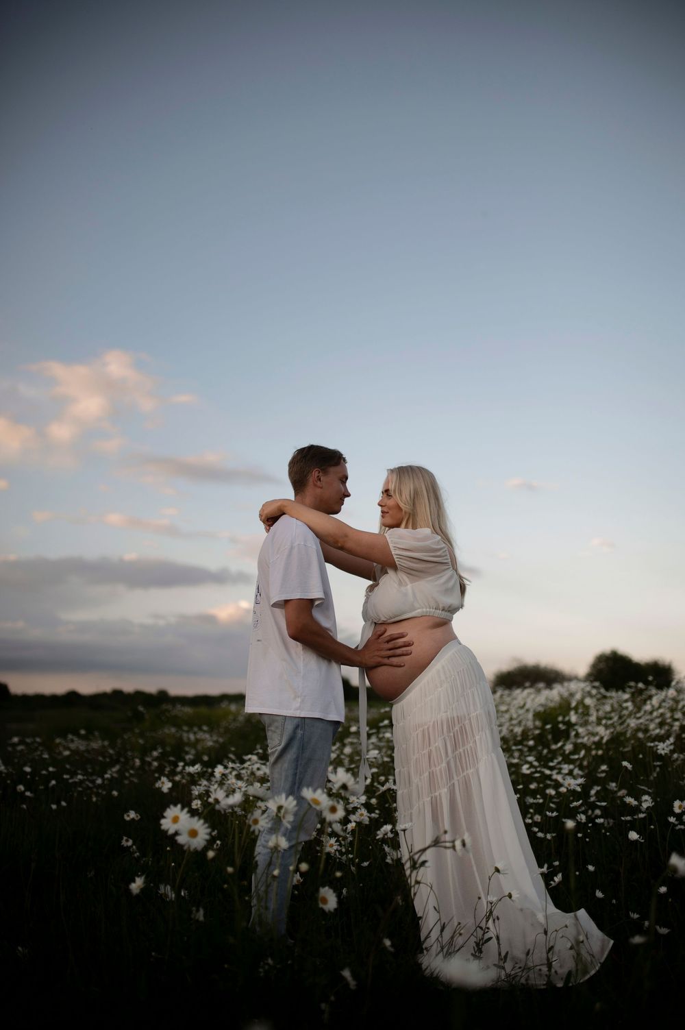 A romantic couple embraces in a field of white wildflowers at sunset with soft clouds in the background.