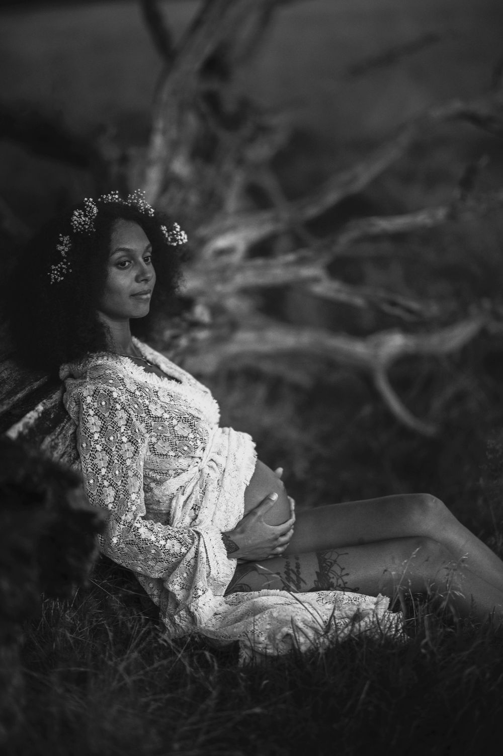 A black and white portrait of an expectant mother in a lace dress with flowers in her curly hair sitting outdoors.