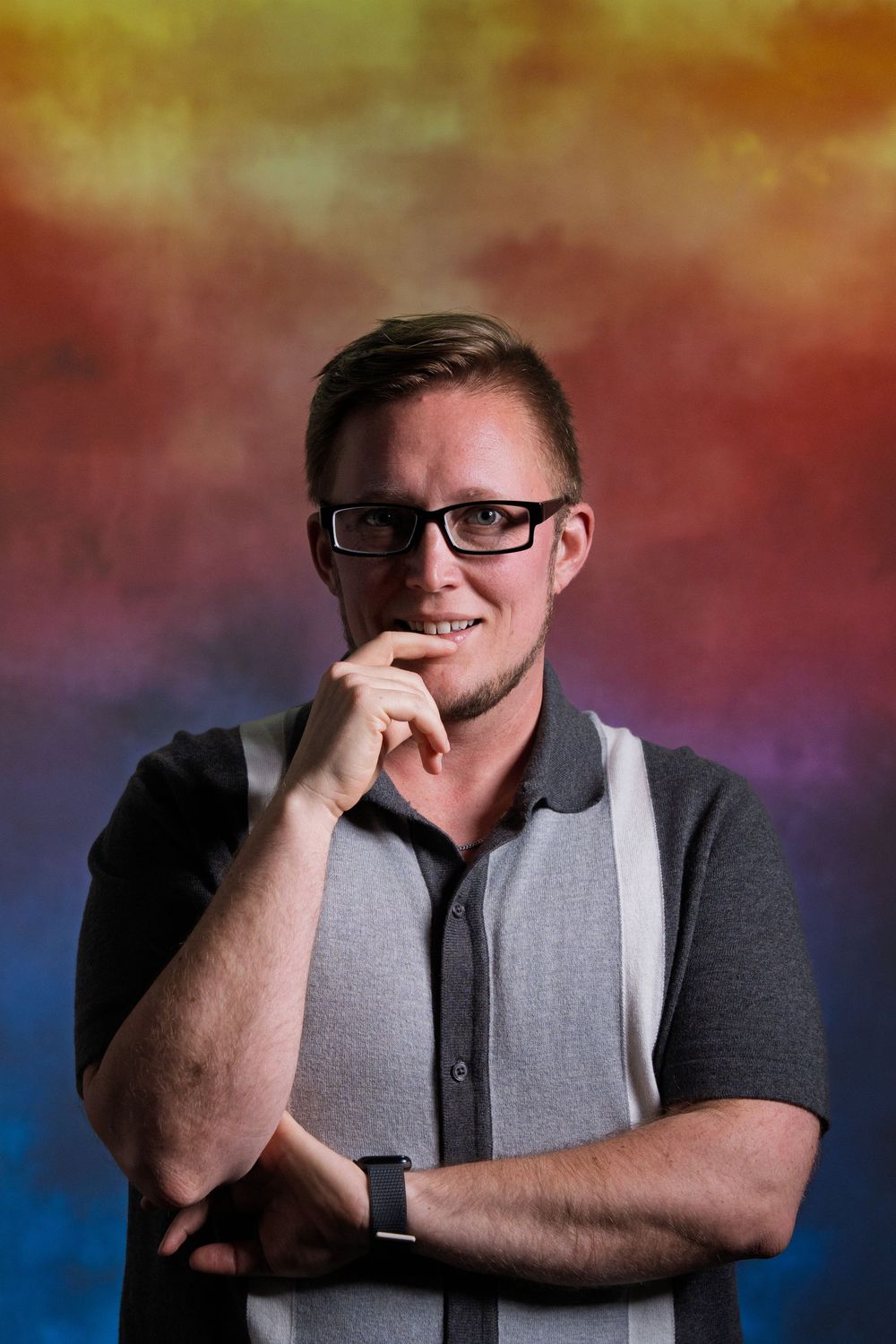 Professional studio portrait of a person in glasses and a grey polo shirt against a colorful gradient background.
