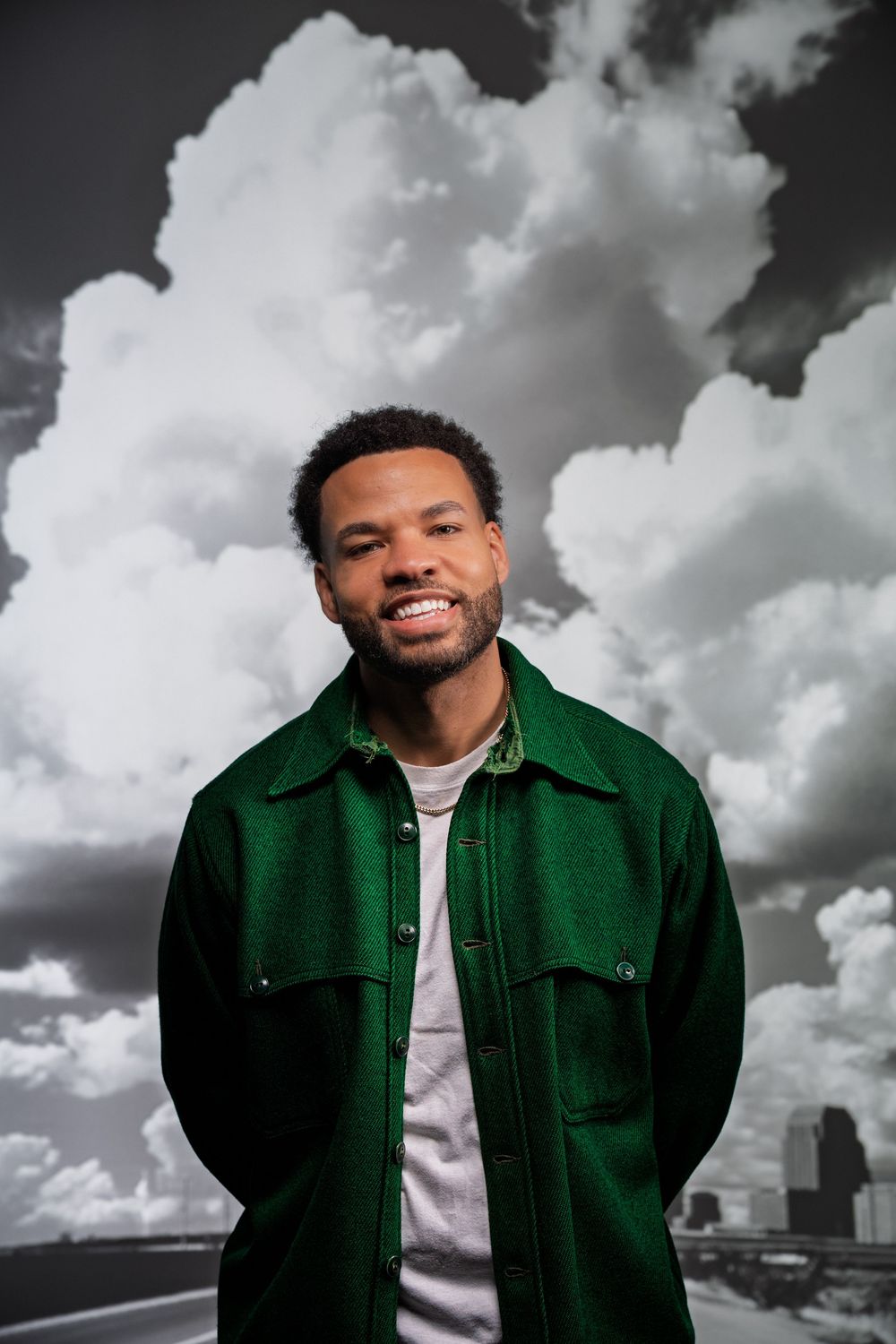 Person in emerald green jacket stands against dramatic cloudy sky with city skyline in background.