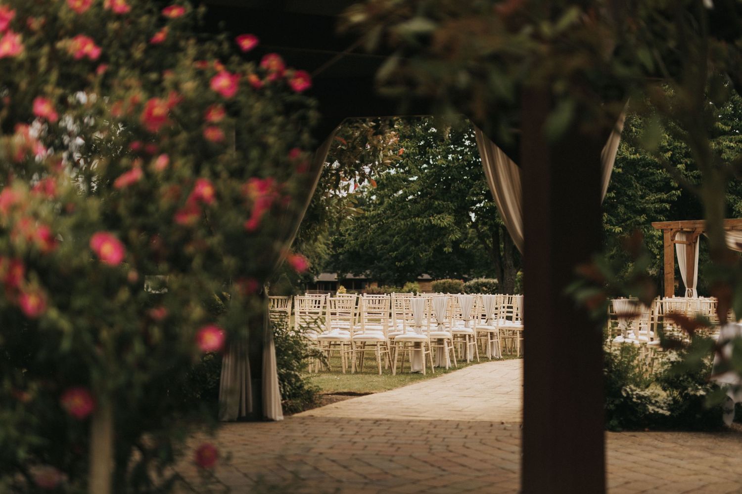 White wedding chairs line a garden path framed by trees and pink roses at an outdoor ceremony venue.