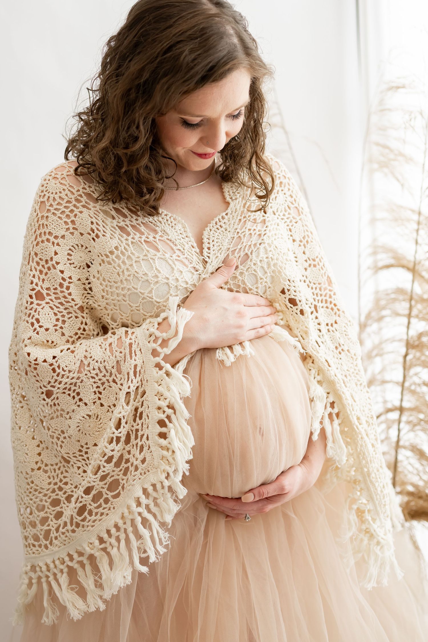 An expectant mother in a cream crochet shawl and blush tulle dress holds her belly by a window with pampas grass.