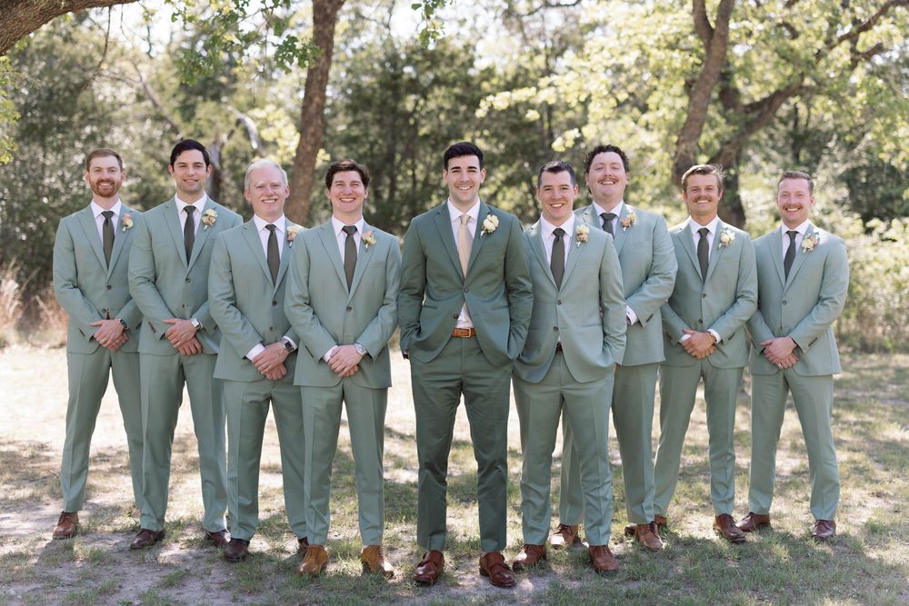 A group of groomsmen wearing matching sage green suits stand together outdoors for a wedding photo.