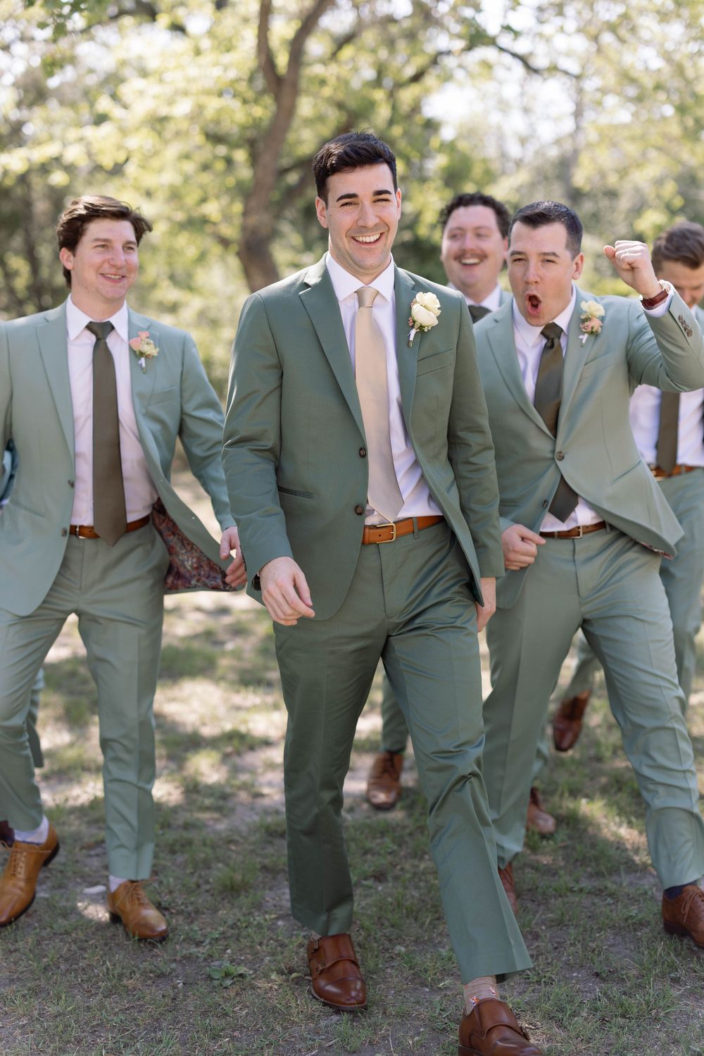 Groomsmen in matching sage green suits and pink ties walk together outdoors while sharing a laugh.
