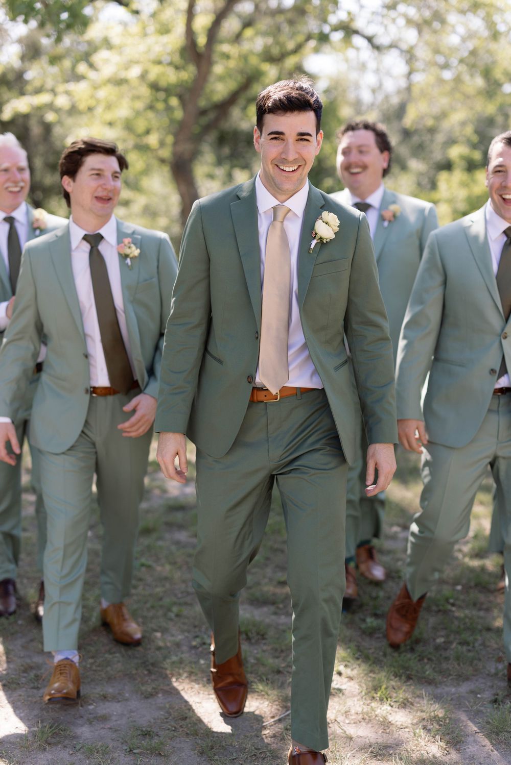 Wedding party wearing sage green suits with brown shoes walking and smiling outdoors in natural lighting.