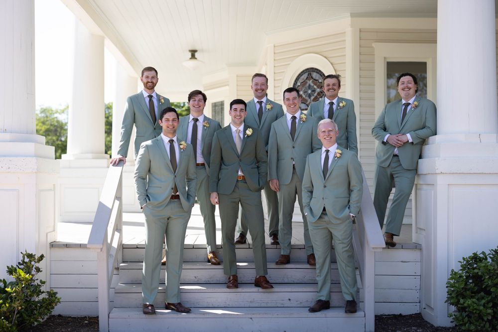 A wedding party wearing sage green suits stands on white steps outside a building with columns.