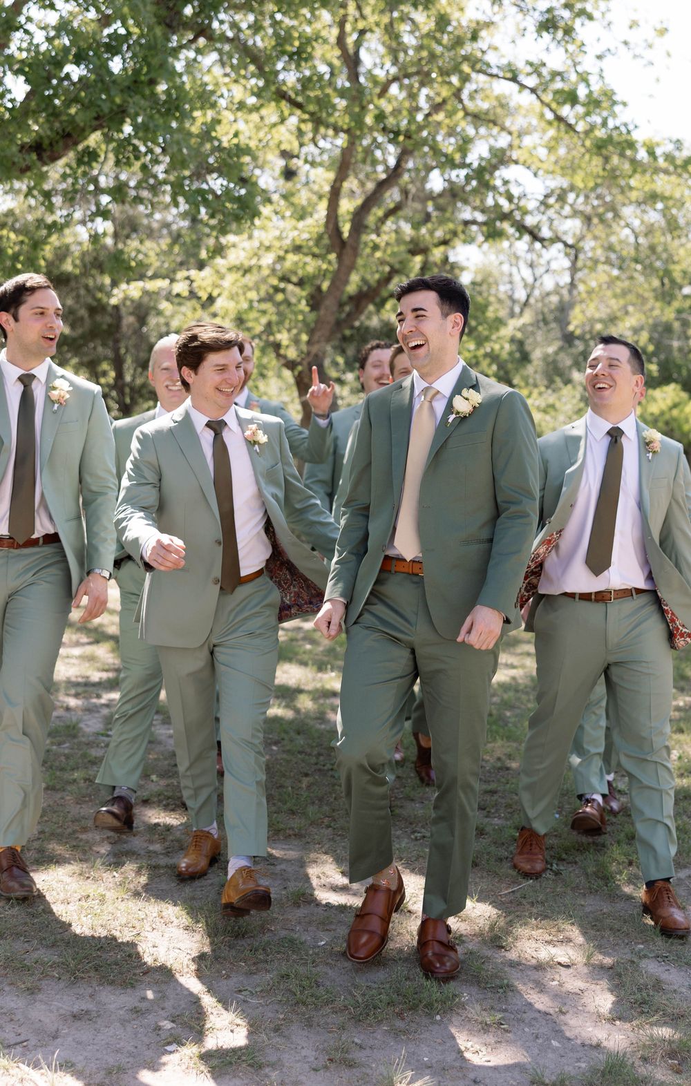 Groomsmen in sage green suits and brown shoes walk together laughing on a sunny outdoor path.