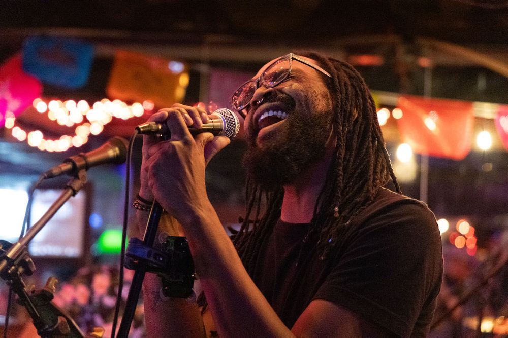 An enthusiastic performer with dreadlocks sings passionately into a microphone on stage with colorful lights in the background