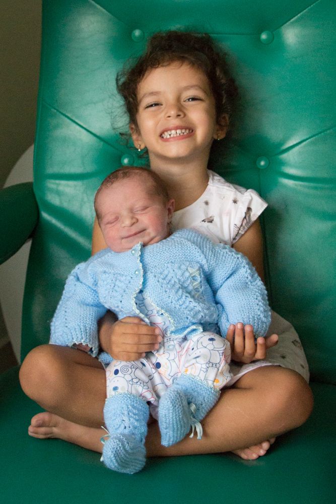 A child in a polka dot dress holds a newborn wearing a blue knitted outfit while sitting in a turquoise chair.