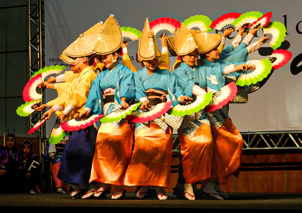 Dancers in vibrant orange dresses perform traditional Japanese folk dance with colorful flowing skirts on stage.