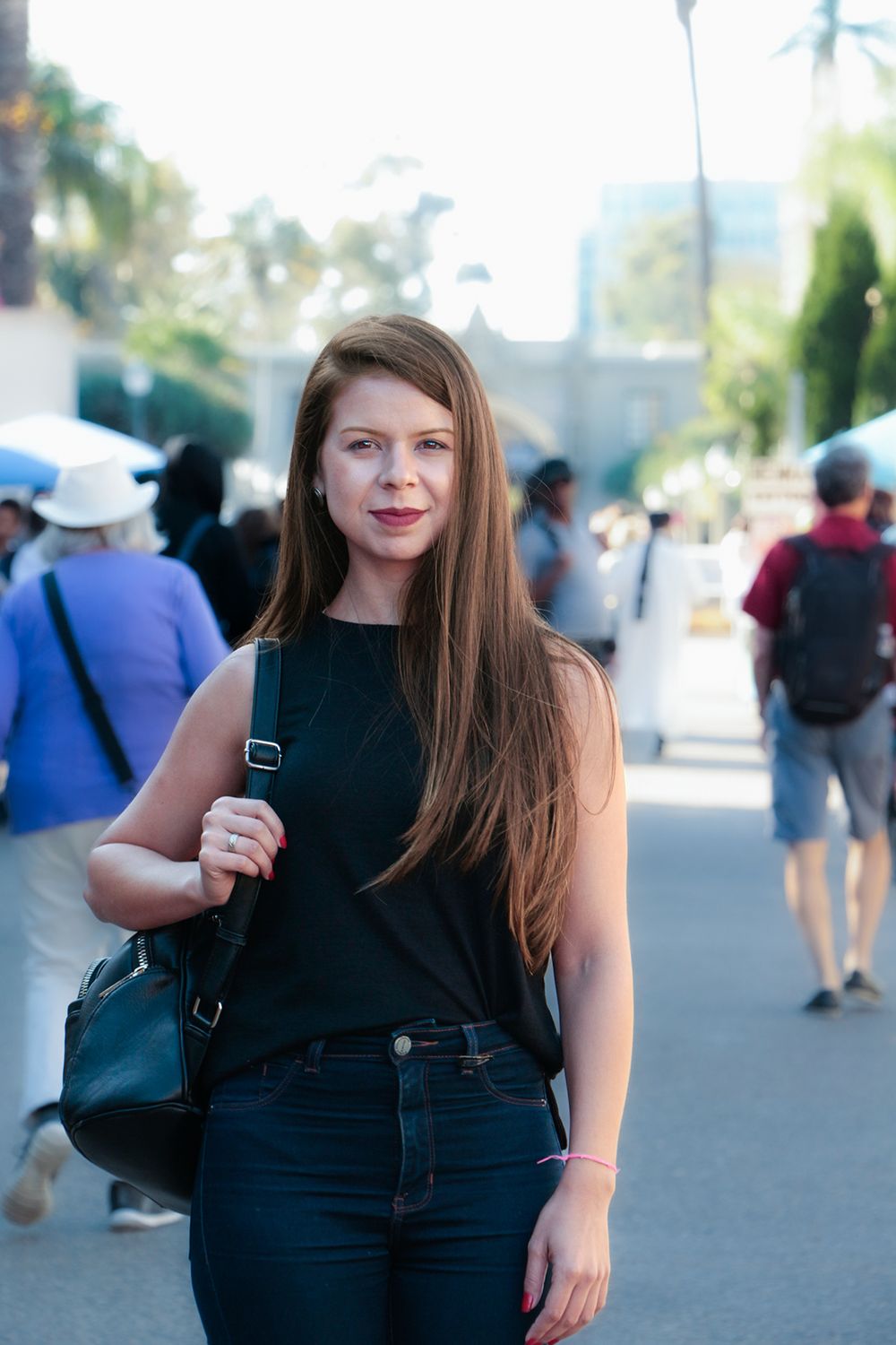 Woman wearing dark sleeveless top and jeans standing outdoors at a crowded tourist location on sunny day.