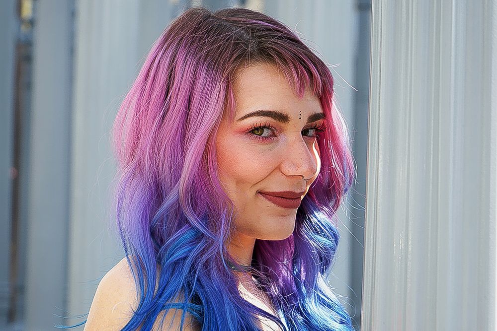 Portrait of person with vibrant pink and blue ombre hair styled in long waves against a light background.