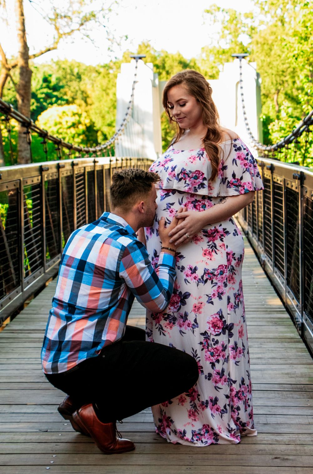 A tender moment captured on a wooden suspension bridge as a couple in summer attire shares a maternity moment photo-shoot.