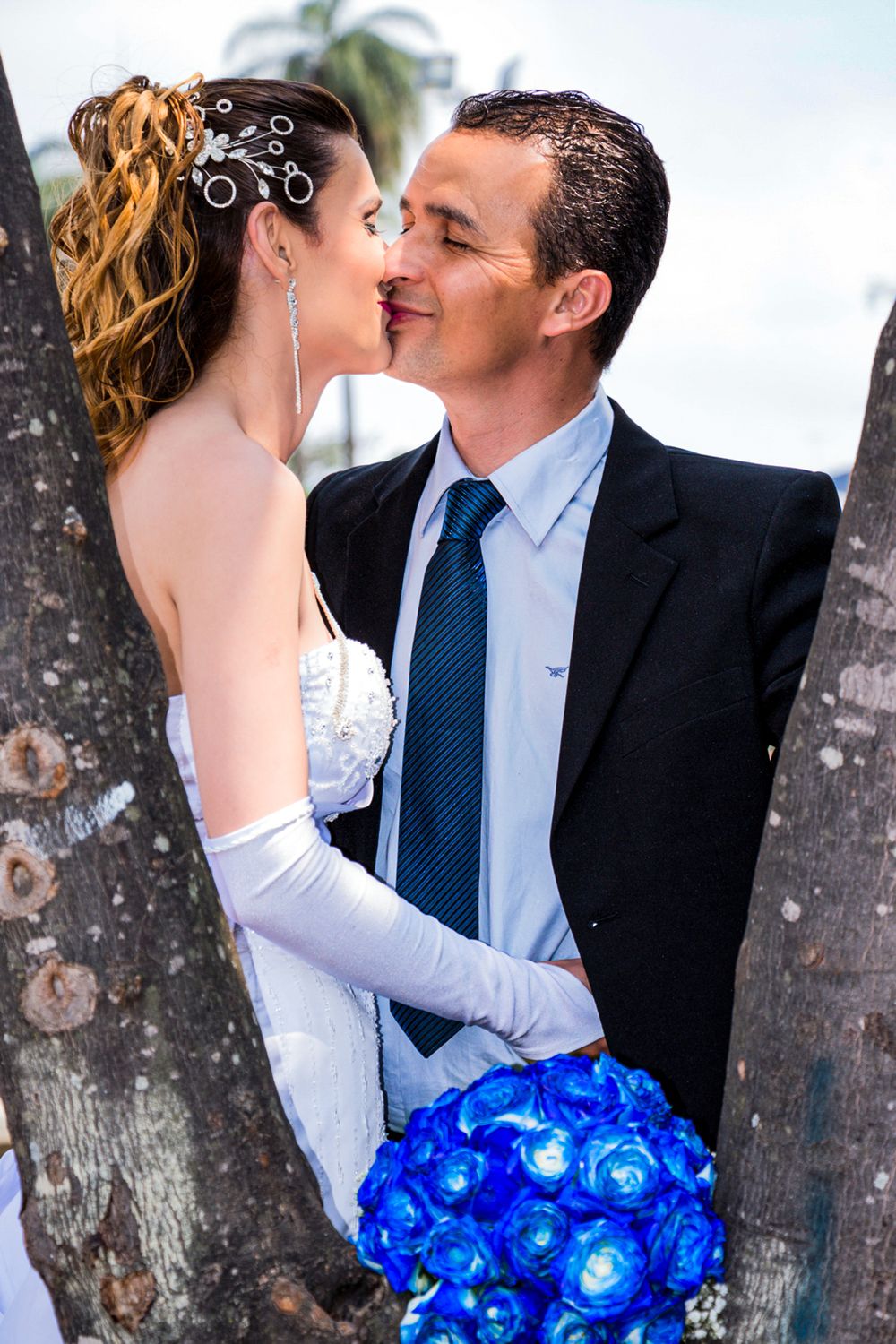 Wedding couple sharing a romantic moment between palm trees with bride holding vibrant blue bouquet during photo-shoot.