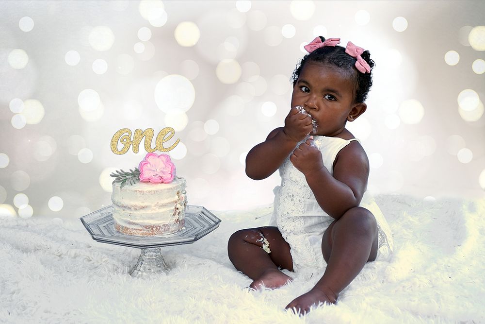 Baby in white dress smashes cake and plays with frosting during festive first birthday cake smash photoshoot.