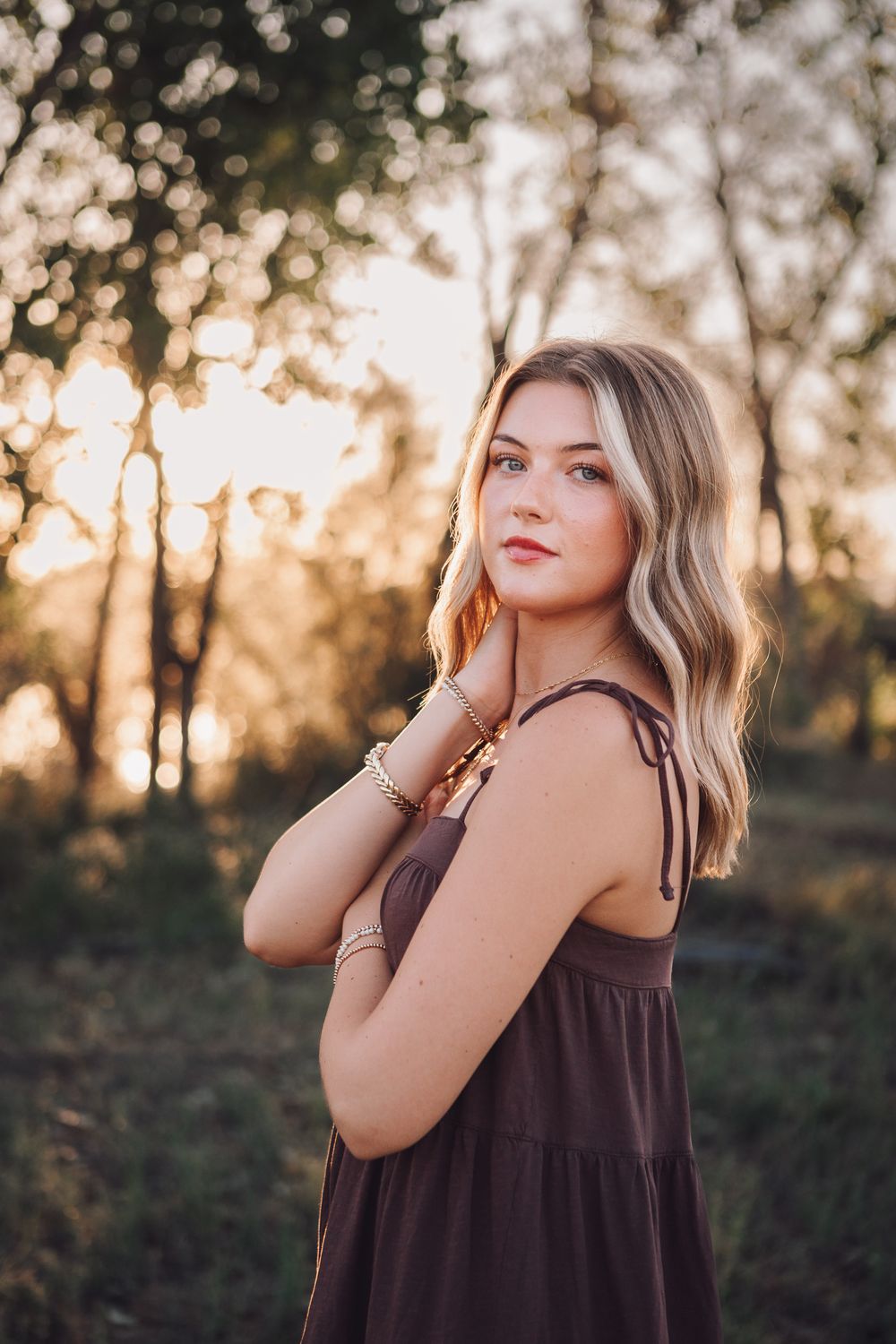 Portrait of person in brown dress with wavy blonde hair standing in nature during golden sunset hour.