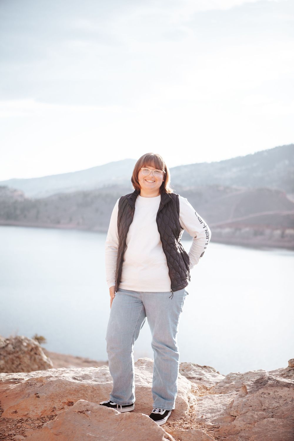 Someone poses confidently on a rocky overlook with a scenic mountain lake view in the background.