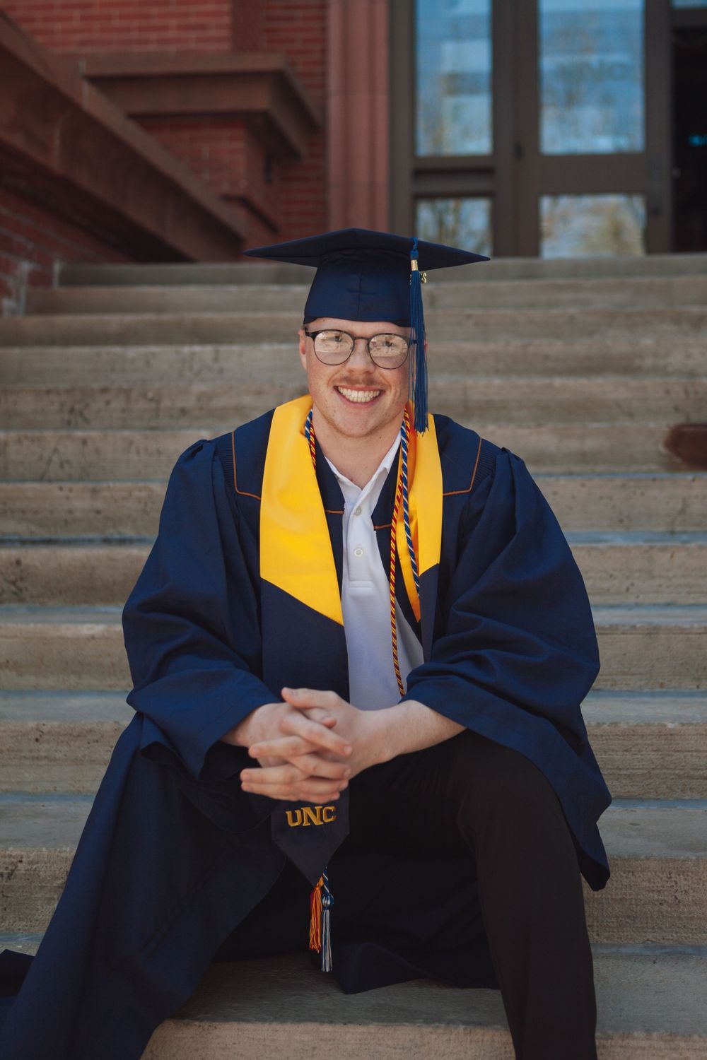 Graduate in cap and gown with gold stole sits smiling on stone steps in front of brick building.