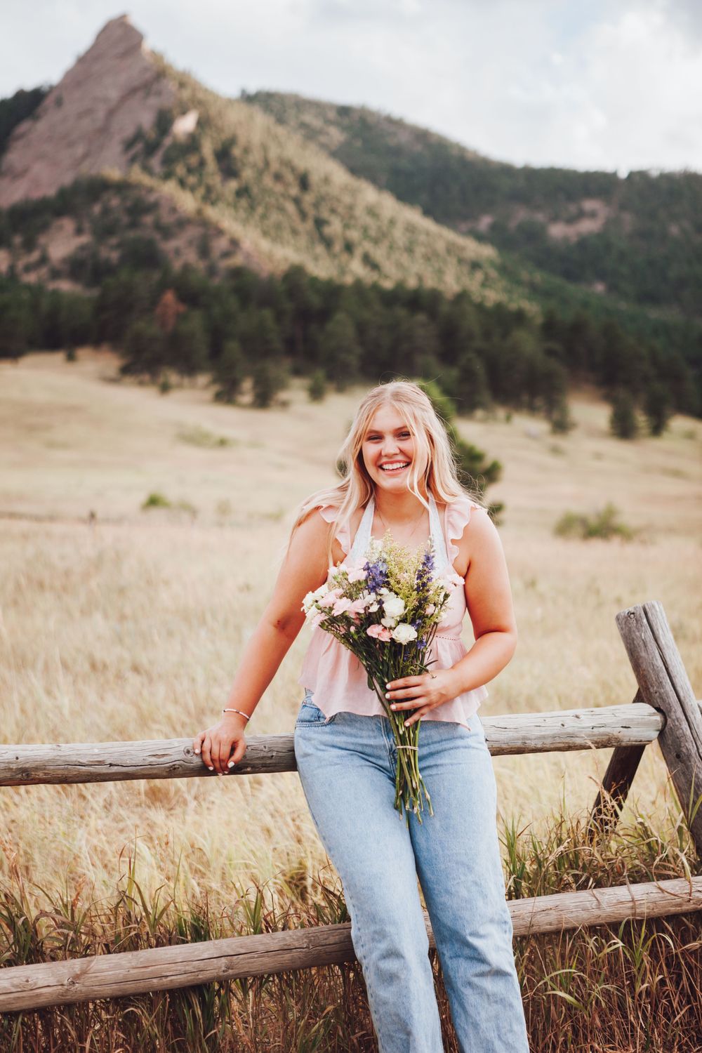 Person sitting on rustic wooden fence in field wearing jeans and holding wildflower bouquet against mountain backdrop.