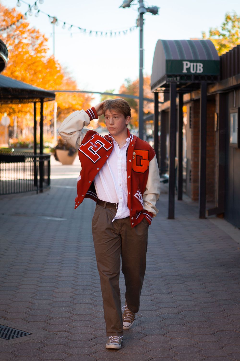 Person in red letterman jacket and brown pants walks along a brick sidewalk with autumn trees and string lights overhead.