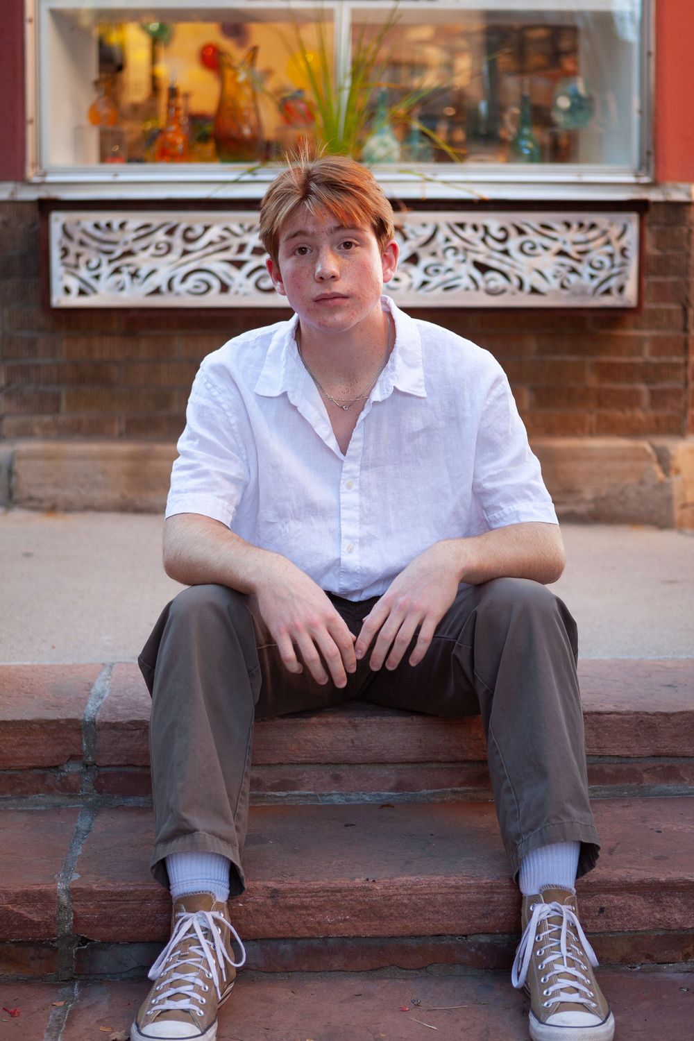 Person in white button-down shirt and gray pants sits on brick steps in front of decorative storefront window.