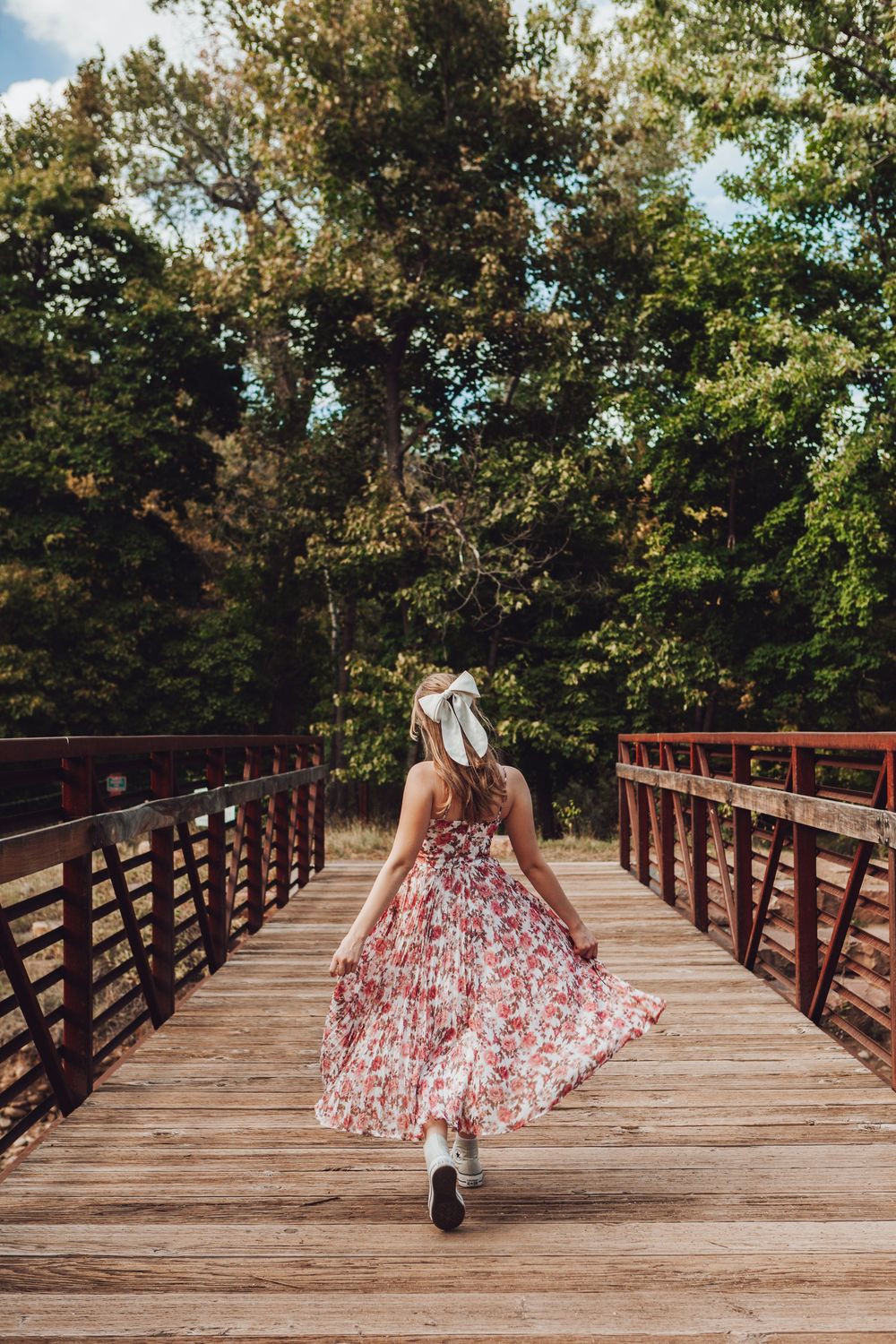 A woman in a floral dress twirls on a wooden bridge surrounded by lush green trees on a sunny day.