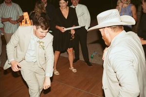 Two people dancing on a dance floor at a party, one in a tan suit and one wearing a white cowboy hat.