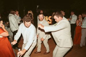 Wedding guests in beige and white suits energetically dance together on the reception dance floor at night.
