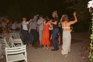Wedding party guests dancing and celebrating outdoors at night with white chairs visible.