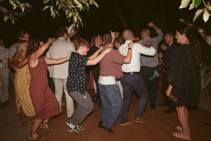 Enthusiastic wedding guests form a dance circle during an outdoor evening reception celebration.