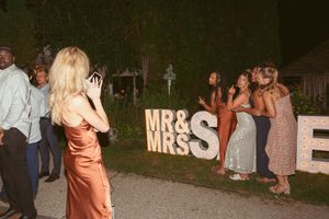 Wedding guests pose for photos near illuminated 'MR & MRS' sign at outdoor reception.