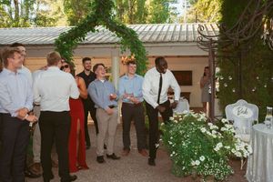 Guests in formal attire gather under a green archway at an outdoor wedding venue.
