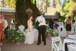Outdoor wedding reception with white chairs and floral decorations under tall evergreen trees.