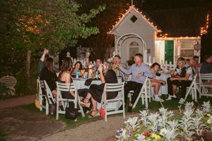 Wedding guests seated at outdoor tables with white chairs under string lights at dusk.