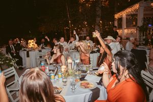 Wedding guests at a lively reception dinner raising their glasses in celebration at round tables with glowing lights.