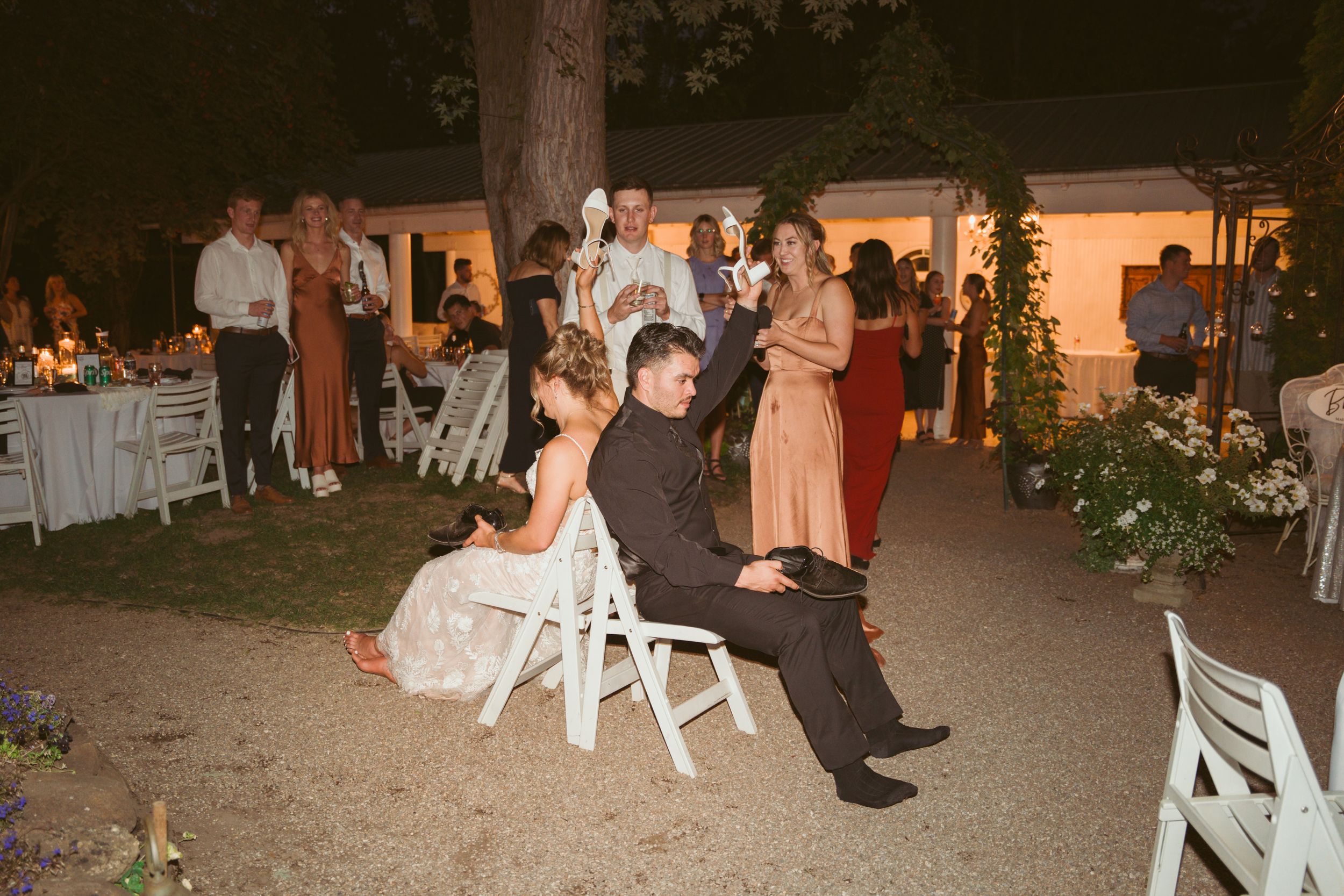 Wedding guests participate in a playful game during an outdoor evening reception with string lights and white chairs.