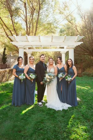 Wedding party in slate blue dresses posing outdoors under white pergola with lush green grass and trees.