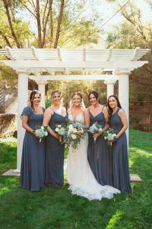 Bridal party in dusty blue dresses standing together under a white pergola in a garden setting.