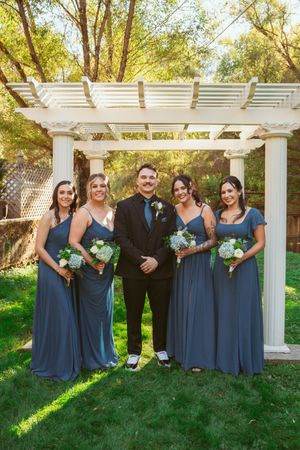 A bridal party stands under a white garden pergola on a sunny day, with everyone wearing navy blue attire.