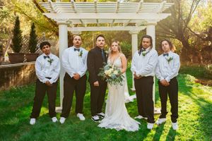 Wedding party poses underneath a white pergola in a garden setting with green grass and sunlit trees.