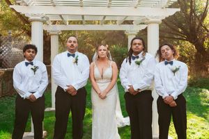 A wedding party wearing formal black and white attire poses together under a white garden pergola on a sunny day.