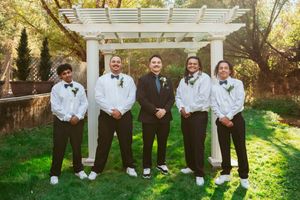 A wedding party in formal attire poses together under a white pergola in a garden setting.