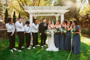 A wedding party in gray dresses and black suits poses together outdoors under natural lighting.