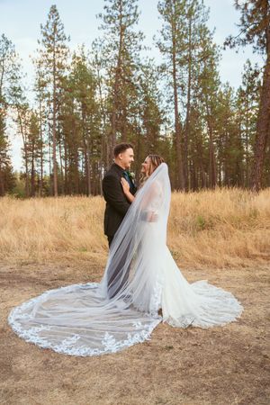 A romantic wedding couple poses in a field of golden grass with tall pine trees in the background at sunset.