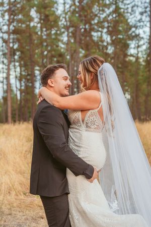 A romantic wedding couple embracing and dancing in a sunlit field surrounded by tall pine trees.