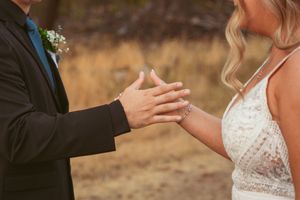 A close up of hands touching between someone wearing a black suit jacket and someone in a white lace wedding dress.